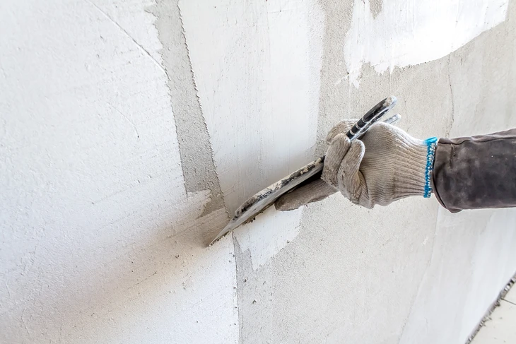 Ouvrier en combinaison blanche et masque respiratoire utilisant une ponceuse électrique murale dans un intérieur en rénovation