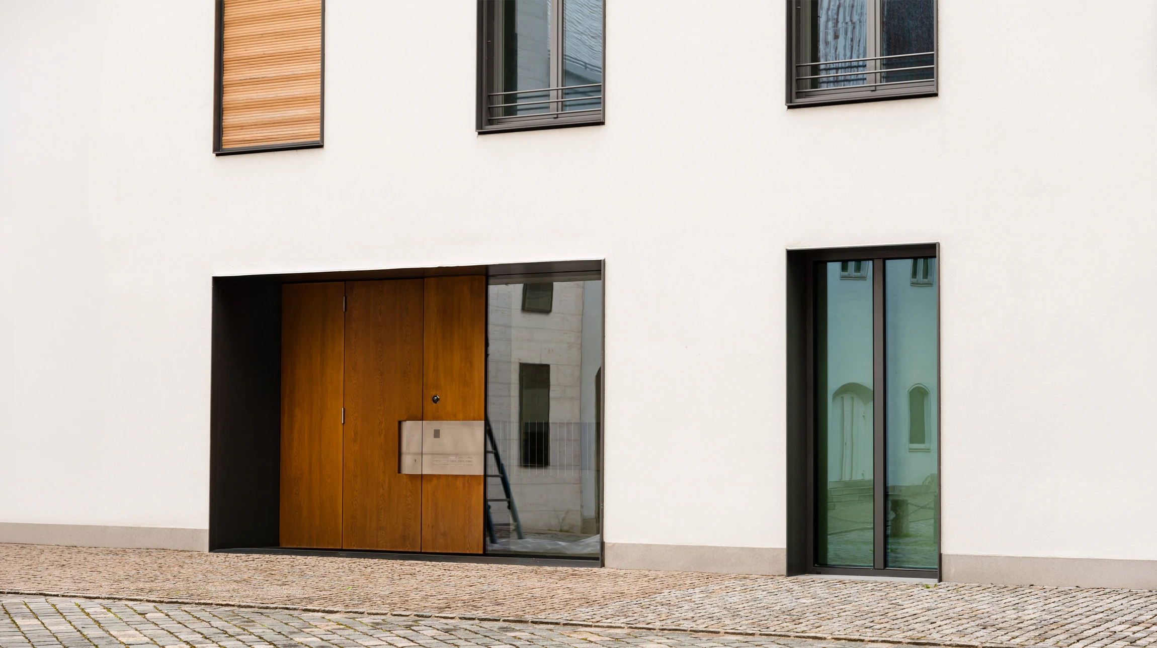 Façade extérieure moderne avec mur en briques peintes vert pâle, porte d'entrée bleue encadrée de blanc, luminaire suspendu doré, plaque d'adresse numérotée, haie verte taillée à la base et trottoir beige