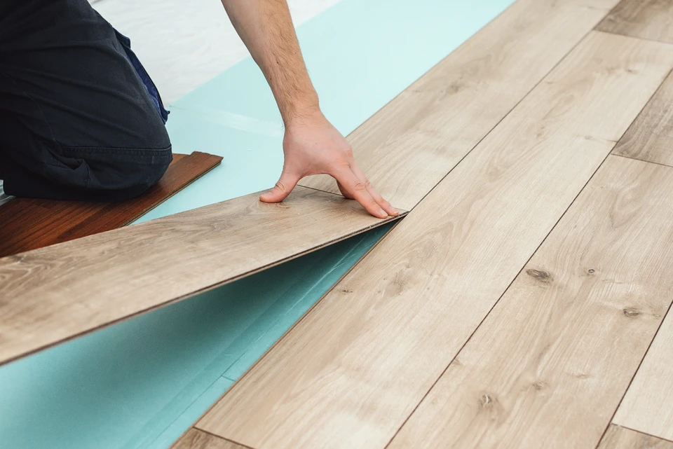 Homme installant un parquet flottant à l'intérieur, agenouillé sur le sol, mesurant une lame avec un mètre ruban jaune
